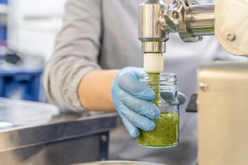 Pesto sauce Industrial process. High quality sauce production. Worker operator at the food factory behind the machine pours pesto sauce into jars. Italian basil taste in Pesto sauce