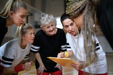 Female sport coach with clipboard discussing tactics with young and old women team training for match in gym.