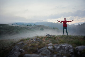 Senior woman doing breathing exercise in nature on early morning with fog and mountains in background.