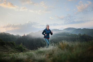 Senior woman jogging in nature on early morning with fog and mountains in background.
