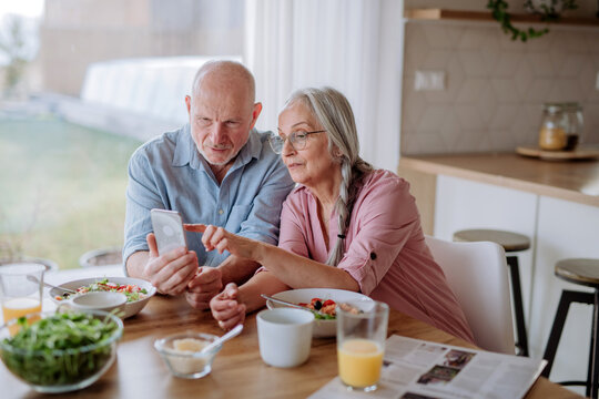Happy senior couple using smartphone during eating dinner together at home. - Powered by Adobe