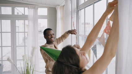 Happy multinational couple hangs colorful pennant banners on panoramic window. Man and woman...