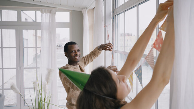 Happy multinational couple hangs colorful pennant banners on panoramic window. Man and woman prepare for birthday party decorating room