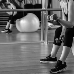 Fototapeta na wymiar Sporty athletic woman lifting dumbbells while sitting on fitball in gym, black and white image