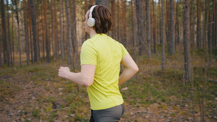 Young man in white wireless headphones runs through pine forest. Runner listens to music during cardio training outdoors