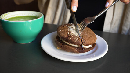 Woman cuts brown cake with stainless steel cutlery in cafe. Plate with dessert and cup of green matcha served on table in cafe closeup