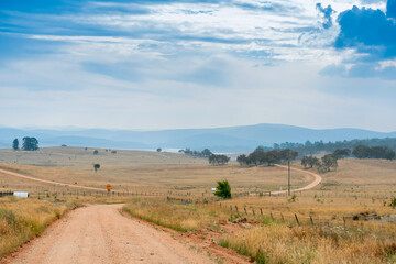 Fototapeta na wymiar Country unsealed gravel dirt road surrounded by farms and fields in Australia. Mountains on the horizon. Road trip rural travel