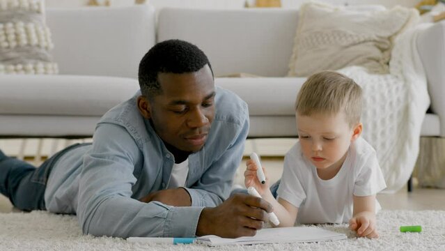 Black African family dad and caucasian son together at home. Young father and little boy smile at camera and lying on warm floor and drawing coloring on book with color pencil crayon at living room.