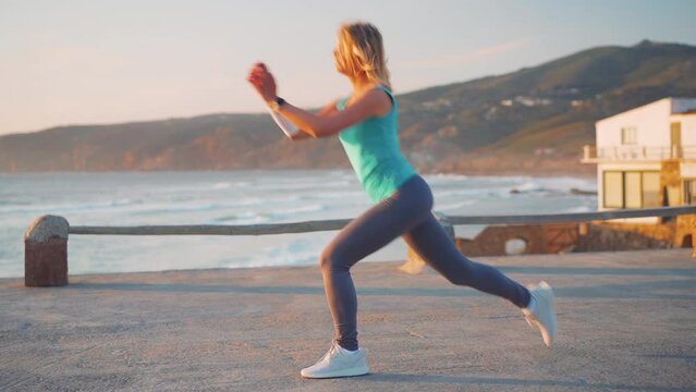 Slow motion shot young female athlete practicing lower body exercise. She doing forward moving lunge against amazing ocean mountain scenery. Caucasian fitness female doing lunges workout training