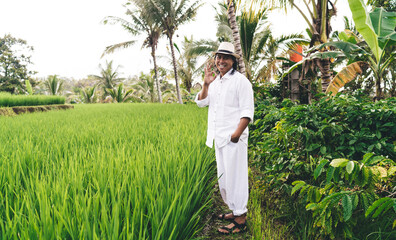 Full length portrait of joyful male farmer in casual clothes showing OK during daytime at rice fields with coffee bush for growing caffeine beans, cheerful Balinese man enjoying agriculture business