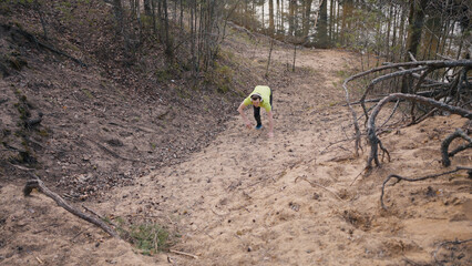 Young man during jog climbs on steep hill without stopping. Runner in bright yellow t-shirt and headphones climbs on sand hill with help of hands