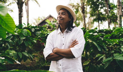 Cheerful male agriculture owner with crossed hands smiling while visiting coffee business during daytime for seedling at tropical plantation of Ecuador, happy man in hat farming at countryside