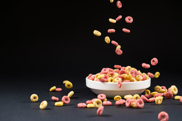 Multicolored breakfast cereal rings are poured into a plate on a black background, a place for text.