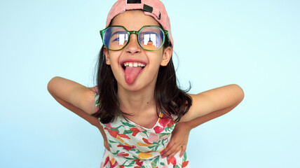 Funny child in eyeglasses and pink cap looking at camera showing her tongue. A kid sticking tongue out posing over studio blue background. Positive little girl making grimace.
