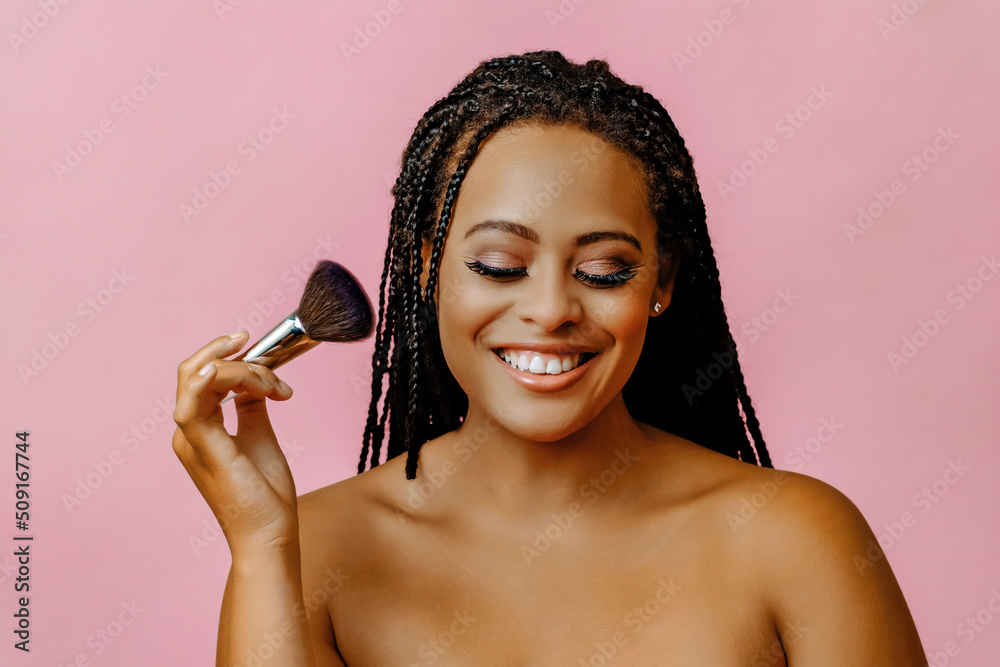 Wall mural happy toothy smile young adult woman with dreadlocks eyes closed on a pink background holding makeup