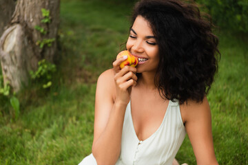 happy and young african american woman in white dress eating peach in park.