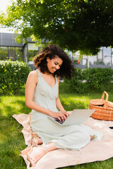 smiling african american woman using laptop near wicker basket on blanket in park.