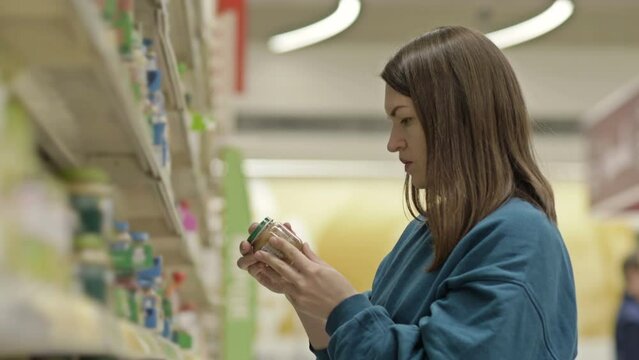 Young Woman Customer Carefully Examines The Label On A Jar Near The Shelves With Baby Food In A Supermarket.