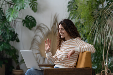 Young hispanic woman waving hello, having video call on laptop sitting in armchair in cozy home garden. Italian female talking in video chat on computer surrounded by houseplants in apartment. 