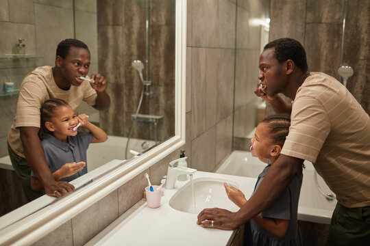 Side view portrait of black father and daughter brushing teeth together in bathroom and looking at mirror, copy space