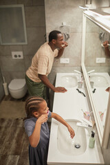 High angle of black father and daughter brushing teeth together and looking at mirror in bathroom