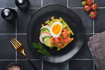 Savory waffles. Breakfast green waffles wild garlic or spinach with egg, tomato, salmon on wooden plate for breakfast on old black tile table background. Light dinner, lunch or breakfast. Top view.