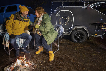 Obraz na płótnie Canvas Mature family at campfire near camper away in nature. Mature couple at campfire romantic dinner, hot natural tea sitting on the plaid blankets in winter relaxation journey. Family journey concept. 