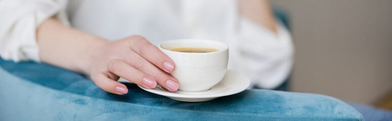 Cropped view of woman holding cup of coffee on saucer on armchair, banner.