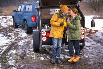 In love couple romantic journey winter camper vacation, man kisses woman while drinking hot tea in cups standing at mini camper back kitchen. Family spend time camping, journey together. 