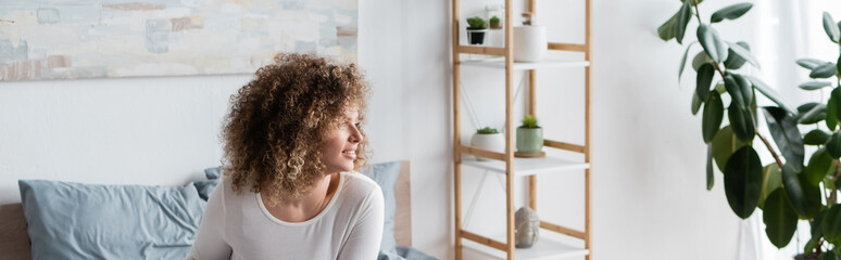 happy woman with curly hair looking away in bedroom near rack and potted plants, banner.