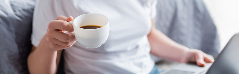 partial view of blurred woman holding cup of coffee at home, banner.