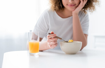 cropped view of smiling woman holding spoon with corn flakes near bowl and glass of orange juice.