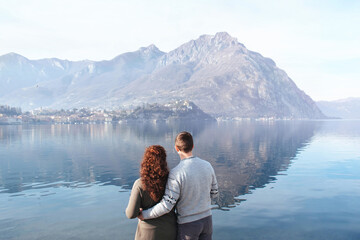 Fototapeta na wymiar Soft focus, back view of couple embracing each other on the shores of Lake Lecco, Italy. People in love on a background of mountains and water outdoors enjoy a date. Romantic relationship. Copy Space
