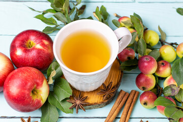 A white mug with tea on a wooden stand, ripe red apples, cinnamon sticks and anise on a blue (mint) background. The composition.