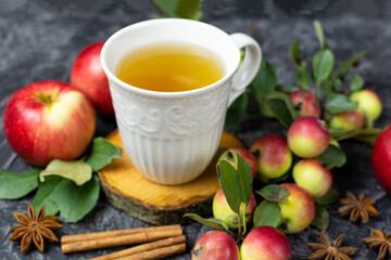 A white mug with tea on a wooden stand, ripe red apples, cinnamon sticks and anise on a black (dark) background. The composition