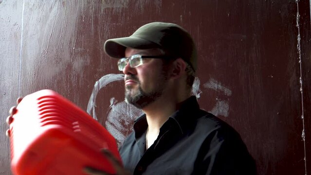 A man with a red canister. Canister for gasoline. Glasses on the face. Bearded man in a baseball cap. Run out of petrol. Garage gate on the background. Portrait to the waist. Close-up.