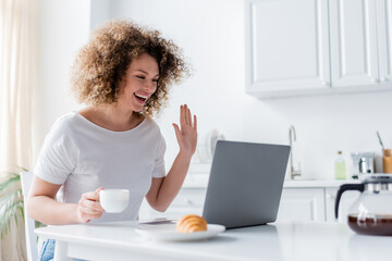 happy woman with coffee cup waving hand near laptop and blurred croissant.