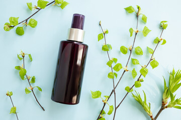 Brown cosmetic spray bottle and birch branches with young small leaves on blue background. Mockup....