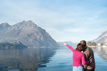 Fototapeta na wymiar Soft focus, back view of couple enjoying a romantic date on the shores of Lake Lecco, Italy. The girl indicates direction with her hand. People in love on background of mountains, water. Copy space