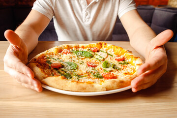 Italian pizza with chicken, cherry tomatoes, mozzarella and parmesan cheese, basil on plate on wooden table and male caucasian hands. Top front view, lifestyle photo, closeup.