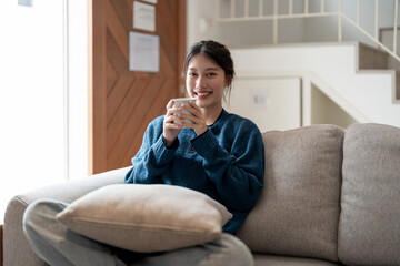 Happy young asian woman sitting on a couch with a cup of coffee at home