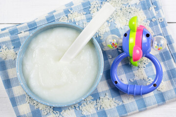 Rice porridge for the baby from ground cereals in a blue bowl with a spoon, a bright rattle toy on a cloth napkin on a white wooden background. Baby's first complementary food, baby nutrition.