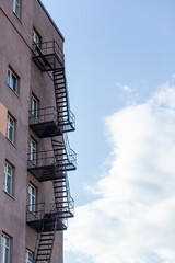 Silhouette of a fire escape on a high-rise building against a blue sky with clouds. Some of the stairs are broken. There is free space for text