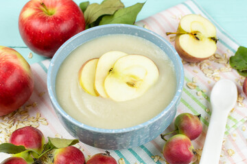 Oatmeal porridge for the baby from ground cereals in a blue bowl with a spoon, red ripe apples on a cloth napkin. The first complementary feeding of the child.
