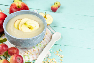 Oatmeal porridge for the baby in a blue bowl, red juicy apples on a blue wooden background. Baby's first complementary food, baby nutrition. Space for text.