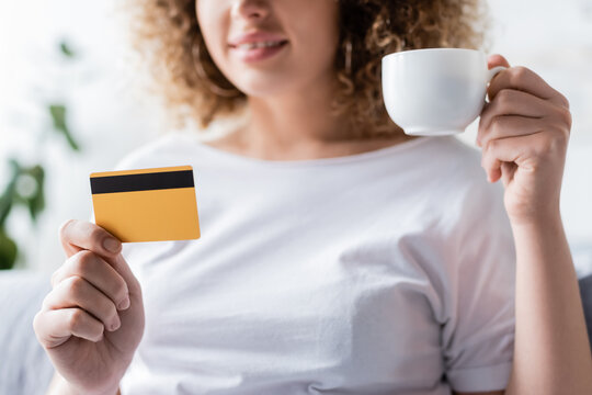 partial view of smiling woman holding coffee cup and credit card on blurred background.