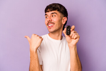 Young hispanic man holding keys car isolated on purple background points with thumb finger away,...