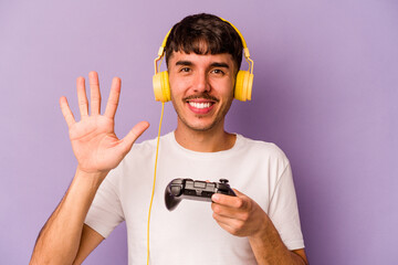 Young hispanic man playing with a video game controller isolated on purple background smiling cheerful showing number five with fingers.