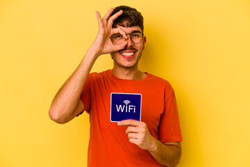 Young hispanic man holding wifi placard isolated on yellow background excited keeping ok gesture on eye.