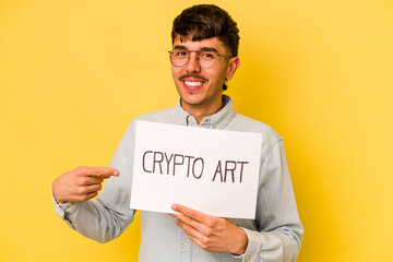 Young hispanic man holding crypto art placard isolated on yellow background smiling and pointing aside, showing something at blank space.